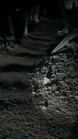 A dark, grainy video captures a rough, uneven dirt path strewn with rubble and debris. The path appears to be within a damaged structure or area, with broken concrete pieces and fragmented building materials scattered about. In the background, indistinct structures and possible makeshift shelters are visible, shrouded in darkness. A string or wire stretches across the path, illuminated by a bright light source that also casts long shadows. Towards the end of the video, the headlights of a vehicle emerge from the darkness, casting a bright beam onto the path and highlighting the debris. The red taillight of the vehicle is also visible. The overall impression is one of devastation and a lack of proper infrastructure, possibly in a conflict zone or disaster-affected area.