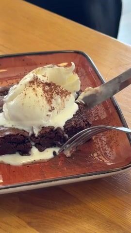 The video shows a close-up of a dessert on a dark orange rectangular plate. The dessert consists of a brownie topped with two scoops of vanilla ice cream, dusted with cocoa powder. A fork is seen lifting a piece of the brownie and ice cream. To the left of the brownie, there are sliced strawberries and a sprig of mint. The plate is on a wooden table, and a knife is also visible on the right side of the plate. In the background, a person in a black shirt is partially visible. The scene appears to be indoors, likely in a restaurant.