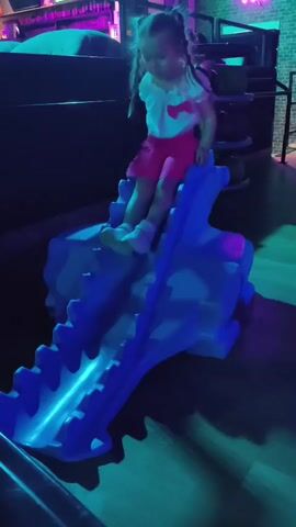 A young girl with blonde pigtails, wearing a white top and pink shorts, sits on top of a blue, dragon-shaped slide with illuminated blue lights. The slide has a toothed texture along its length. The background is dark with purple and teal lighting, suggesting an indoor environment like an arcade or play area. The girl is looking down towards the slide, and her right hand is extended down.