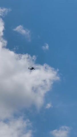 A drone is captured in flight against a clear blue sky with scattered white clouds. The drone appears to be small and white with dark propellers. It is positioned in the center of the frame, slightly to the left, and is the sole subject of the image. The background is a vast expanse of sky, suggesting an outdoor setting during the daytime. The lighting indicates it is a bright, sunny day, possibly mid-morning or afternoon. The presence of clouds suggests fair weather conditions. There are no visible people, and no text is present in the media file. The overall impression is one of technological advancement and aerial observation.