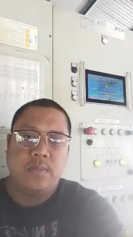 A man wearing glasses and a black t-shirt looks into the camera. He is standing in front of a control panel with a screen and several buttons and switches. The control panel appears to be part of an industrial setting, with a corrugated metal ceiling visible in the background. The man's expression is neutral. There are no other people visible in the video. The lighting is bright, suggesting the video was filmed indoors during the day.