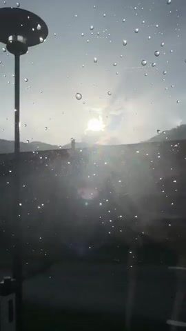 The video shows a view through a window covered in raindrops, with the sun partially obscured by clouds. In the first segment, the sun shines brightly over distant hills. The second segment shows a tree in silhouette against the sun, with a building and green grass in the background. The third segment depicts a dark-colored roof with several dormer windows, with the sky and faint sunlight in the distance. The fourth segment offers a closer view of a white building with a dark roof, a chimney, and a streetlamp, all seen through the raindrops. The final segment returns to the view of the sun over the hills, with the raindrops still visible on the window. The video appears to be shot in a natural, possibly rural, environment.