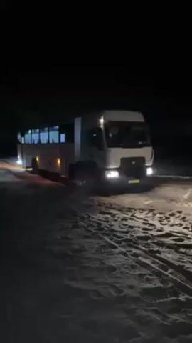 At night, a white bus or truck hybrid vehicle is parked on a sandy surface. The vehicle is illuminated by its headlights and interior lights. Several individuals, who appear to be wearing military fatigues, exit the vehicle onto the sand. The scene is dark with limited visibility beyond the immediate area around the vehicle.