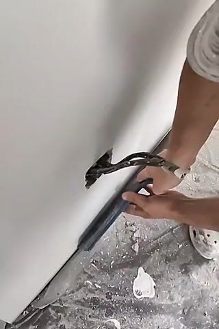 The video shows a close-up of a person's hands working on a white wall. The person is holding a tool, which appears to be a putty knife or a drywall knife, and is scraping at a hole in the wall. There are wires protruding from the hole. The floor around the wall is covered in debris, suggesting construction or renovation work. The lighting is bright and appears to be artificial.