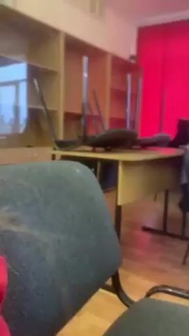 The video shows an interior view of what appears to be a classroom. There are students sitting at desks. Some are facing forward and others are turned towards each other. Red curtains are visible. The room has wooden floors. There is a bookshelf against the wall. Some chairs are on top of desks. There is a notebook and pen on a desk in the foreground. The image quality is poor.