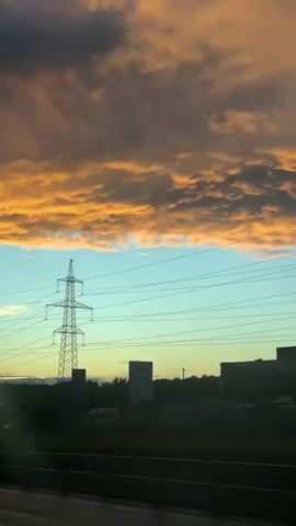 The camera moves forward along a road, capturing the view from a vehicle's window. The sky is divided horizontally into a layer of orange and grey clouds above and a light blue layer beneath. Silhouetted objects dominate the lower half of the frame. These include utility poles, power lines, a billboard advertising "SUNLIGHT", a road sign, and a tall, cylindrical building with antennas. The foreground is blurred, suggesting movement.
