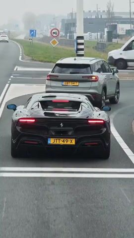 The video shows the rear of a black Ferrari 296 GTB coupe, with Dutch license plate JTT-49-X, as it sits at a traffic light. Other vehicles are visible in the background, including a white van and a grey SUV. The location appears to be a commercial or industrial area with modern buildings. The Ferrari turns right as the red arrow turns green. The background vehicles continue to move through the frame as the Ferrari proceeds.