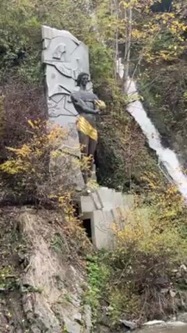 The video shows a statue next to a waterfall in a forest. The statue is of a naked man, draped in a golden cloth, holding something in his hands. It is placed in front of a large stone backdrop with abstract patterns. The waterfall rushes down the hillside, surrounded by lush trees and colorful autumn foliage. The camera pans from the statue to the waterfall and up the side of the hill. The sky is overcast, and the overall scene is natural and scenic.