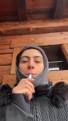 A young woman in a black winter coat with a fur-lined hood and a gray scarf wrapped around her head and neck is standing outside a wooden cabin. She is holding a vape pen to her mouth and appears to be speaking to the camera. Snow is falling lightly around her. The camera moves slightly as she talks.