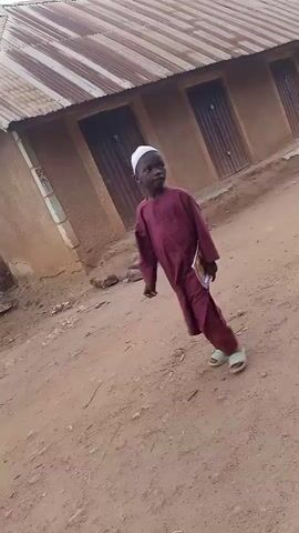 This video captures a moment in Kagarko South, Nigeria, showing a young boy walking forward while holding a book. He is dressed in a traditional maroon tunic and trousers, topped with a white kufi cap, and wears light-colored sandals. The setting appears to be an outdoor area with a dirt ground. Behind the boy, a humble building with a corrugated metal roof and wooden doors is visible. To the right of the building, a motorcycle is parked. The overall atmosphere suggests a typical day in a rural or semi-urban Nigerian community. There is no discernible text in the video. The lighting suggests it could be daytime, with no strong indications of the specific time or weather.