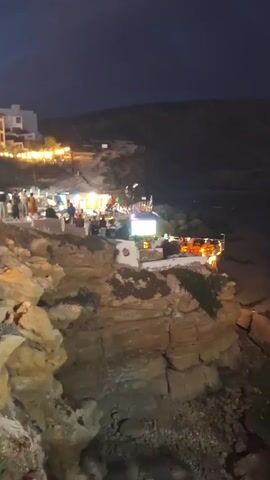 The video captures a nighttime scene on a cliff overlooking a beach. Several buildings with bright lights are visible on the cliff, appearing to be restaurants or bars. People are gathered at these establishments, some standing and others seated at tables. The lighting from the buildings illuminates the area, creating a lively atmosphere. The dark sky and the lights reflecting on the water indicate it is nighttime. The cliff face is rocky and rugged, with some vegetation. The ocean is visible in the background, with gentle waves lapping at the shore. The overall scene suggests a coastal town or resort area enjoying a vibrant evening.