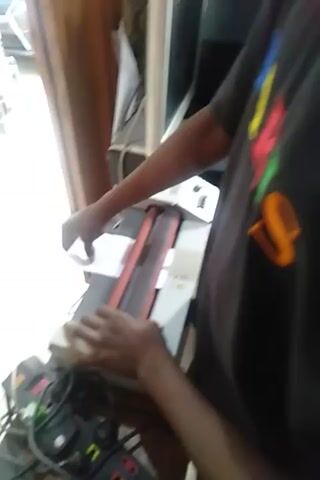 The video shows a person's hands operating a laminating machine. The person is feeding a sheet of white paper into the machine, which has orange rollers. The machine is white with some red accents and a control panel with buttons and a knob. The background is blurred, but appears to be an indoor setting with a desk or table, a monitor, and some wires. The person's hands are dark-skinned. The paper is being fed into the machine from the front.