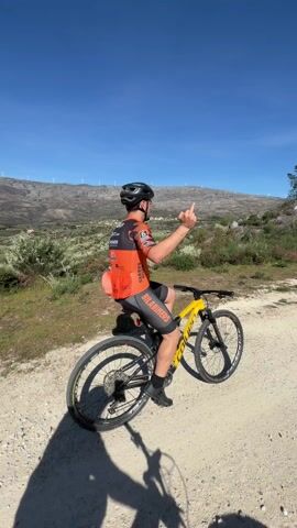 The video shows a man on a yellow and black mountain bike, sitting on a dirt road. He is wearing a black helmet, an orange and black biking jersey, and black shorts. He is giving the middle finger with his right hand. Behind him, there is a mountain range with wind turbines on top of it. The sky is clear and blue. The sun is shining, casting a shadow of the biker on the ground.