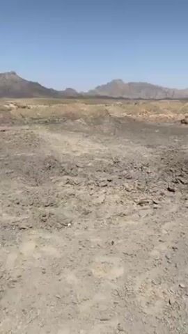 The video shows a barren, rocky landscape with mountains in the distance under a clear blue sky. The camera pans to reveal a partially destroyed stone wall and a pile of rubble. In the background, there is a two-story building with several windows, some of which appear to be broken. The building is a light tan color. The scene suggests damage from some kind of impact or demolition. There are no people visible in the video.