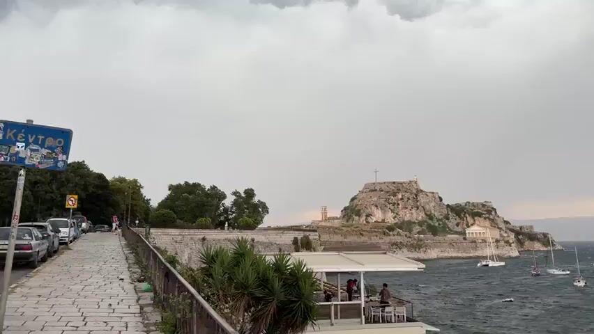 A panoramic video shows a cloudy sky over a choppy sea with numerous sailboats and yachts. On the left, a cobblestone path leads to a stone wall bordering a grassy area and a cluster of trees. Beyond the wall, a rocky fortress with a prominent cross on top is visible, along with a clock tower and some buildings. In the foreground, a white, covered outdoor seating area with tables and chairs is set up, and a few people are seated. The water is dark and wavy, suggesting strong winds or an approaching storm. The overall scene is coastal and appears to be a harbor or bay.