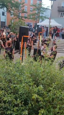 The video captures an outdoor gathering in what appears to be a public square or plaza. A band is performing on a paved area, with several large speakers set up on orange stands. The band members are visible, including a singer with a microphone, a guitarist, and a drummer. A crowd of people is gathered in front of the stage, watching the performance. The audience members are diverse in age and appearance, many dressed in casual or alternative clothing. Behind the performance area, there are buildings, trees, and some temporary tents or canopies, suggesting a possible festival or event. The overall atmosphere seems lively and engaged.