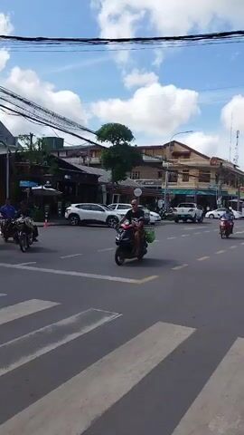 The video shows a busy street intersection with several people on motorcycles and a few cars. The street is paved with asphalt and has white pedestrian crossing lines. On the left side of the frame, there are buildings with storefronts, one of which appears to be a cafe. There are also many power lines overhead. In the foreground, a man on a red motorcycle is riding across the pedestrian crossing. Behind him, other motorcycles and a small white vehicle are also visible. The sky is partly cloudy.