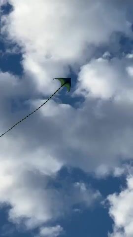 A green and black diamond-shaped kite with a long, striped tail is seen flying in a blue sky filled with white, puffy clouds. The kite is positioned in the upper left quadrant of the frame, with its tail extending diagonally downwards towards the left. The camera is looking up at the kite and sky, suggesting the video was filmed from the ground. The lighting indicates it is daytime.