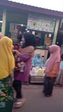 The video shows a busy street market scene. In the foreground, a person in a pink hijab walks away from the camera. The background reveals several market stalls with people browsing and interacting. Some stalls appear to be selling food or produce. Several individuals are wearing hijabs, and some are carrying bags. The environment looks like an outdoor market with temporary structures. The footage is shaky and somewhat blurry, suggesting it was captured on a mobile device.