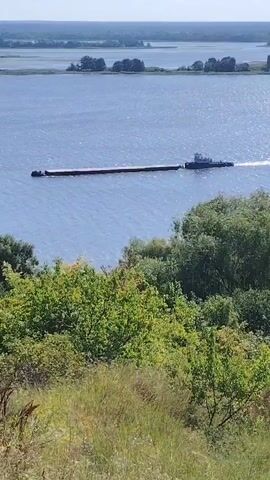 A barge is being towed by a tugboat across a wide body of water. The barge is long and rectangular, and appears to be carrying cargo. The tugboat is smaller and is positioned behind the barge, connected by a tow line. The water is calm, with some ripples indicating movement. In the foreground, there is a bank covered with green trees and grass. In the background, there is a distant shoreline with more trees and vegetation. The sky is clear and blue, suggesting a sunny day. The video is short and shows the barge and tugboat moving steadily across the water.