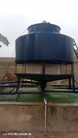 The video shows a large, dark blue cylindrical cooling tower. Water is being sprayed from the top of the tower and falling down through a mesh-like material in the middle section. The water collects in a large, rectangular concrete basin below. The cooling tower is supported by a dark metal frame. There is a ladder on the right side of the tower, leading up to its top. In the background, there are some buildings made of concrete blocks. The ground appears to be dirt and gravel. The sky is overcast.