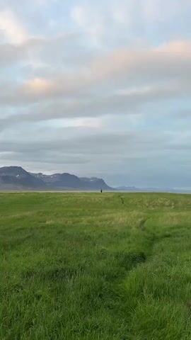 The video presents a serene and expansive landscape, characteristic of Búðir, Iceland. It opens with a wide shot of a vast, vibrant green field, where a narrow path has been naturally worn into the tall grass, leading towards the horizon. In the distance, a solitary figure, seen from behind, walks along this path. Beyond the field, a tranquil body of water meets a dramatic backdrop of rugged mountains, some of which feature prominent patches of snow. The sky is partly cloudy, displaying a mix of soft grey tones and lighter, possibly sun-kissed, areas, indicative of either early morning or late afternoon.

As the video progresses, it slowly zooms in on the person, making them more discernible. They are dressed in dark clothing and appear to be carrying a backpack, reinforcing the impression of a hiker or a leisurely walker. The mountains, with their steep, dark slopes and contrasting white snow, become more defined, highlighting the region's raw natural beauty. The activity shown is a peaceful journey through this grand natural setting. The overall mood is one of quiet contemplation and appreciation for the vastness and majesty of the Icelandic environment. No text is visible in the video.