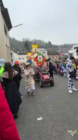 The video shows a carnival parade moving down a paved street through a small town. People in various costumes line both sides of the street and some are walking in the street among the parade participants. A large float shaped like a clown is being pulled by a small red tractor. The driver of the tractor is wearing a green hat and a gray jacket. A woman in a straw hat and beige dress walks on the right side of the street, near the parade. There is debris on the street, including scattered pieces of paper and litter. The buildings on either side of the street are two and three stories tall, and a wooded hillside rises in the background. The sky is overcast and the light is muted.