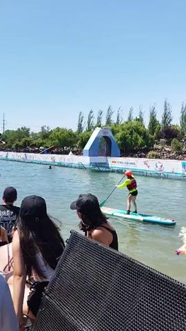 The video shows a crowd of people watching a paddleboard competition on a sunny day. Two paddleboarders, one in green and one in red, are seen in the water competing. The course is marked with a Red Bull archway. The crowd appears to be watching from a raised platform or seating area. There's music playing in the background.