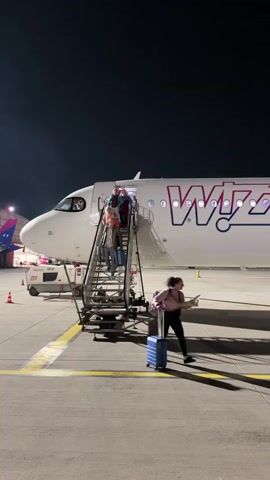 The video shows a Wizz Air plane at night, with people deplaning using a mobile staircase. A woman walks away from the plane pulling a small blue suitcase and holding a piece of paper or ticket. The plane is white with purple and blue markings. The night sky is clear and dark. Other airport equipment is visible in the background.