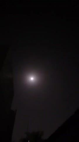 The video shows a night sky with a bright moon. Silhouettes of buildings and trees are visible in the foreground, creating a dark, atmospheric setting. The lighting suggests it is nighttime, with the moon being the primary light source.