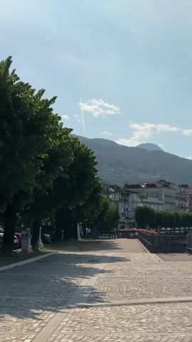 The video shows a scenic view of a lakeside town with mountains in the background. The scene begins with a paved walkway lined with trees and buildings. The camera pans to the right, revealing a wider view of the lake and the town, with boats on the water. The sky is blue with some clouds. A banner on a lamppost reads "stresa festival". The video ends with a view of the lake and the mountains across the water. The overall atmosphere is peaceful and idyllic.
