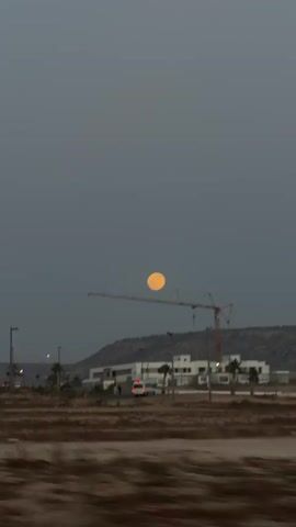 The video shows a landscape at dusk, likely a coastal area, with a large, bright moon visible in the sky. In the foreground, there's a sandy, somewhat barren landscape. In the midground, there are several modern, white buildings under construction, a construction crane, palm trees, streetlights, and vehicular traffic including a white van. The background consists of a low, dark hill. The overall mood is calm, and the lighting is dim due to the time of day. The camera appears to be moving, possibly from a vehicle.