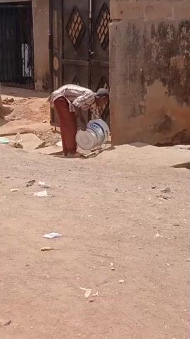 A person is shown outside on a dirt road by a building. The person, wearing a long-sleeved shirt and red pants, is bent over next to a metal drum. The person seems to be adjusting the drum, tilting it slightly towards a wall. The road is littered with trash. In the background, there is a gate with a patterned design. The scene is shot in daylight.