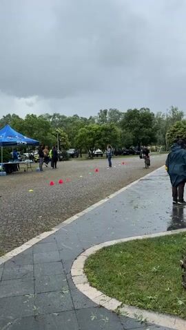 The video captures a rainy outdoor scene where a cyclist is maneuvering through a course marked by orange and yellow cones. The cyclist, dressed in black cycling gear, pedals along a cobblestone path. To the left, a blue tent is set up, with several individuals gathered around a table. Additional people are standing in the background, and some are wearing rain gear. A paved walkway runs alongside the course, and a person in a green raincoat walks along it. The sky is overcast, and there are trees visible in the distance. The ground is wet and scattered with leaves and twigs, indicating recent rain or wind.