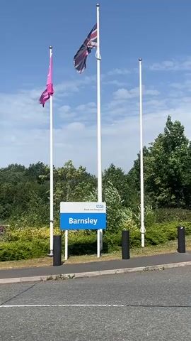 The media file shows a sign for "Barnsley" with the NHS logo, indicating it is likely a public facility in Barnsley, United Kingdom. Three flagpoles stand behind the sign, two of which are flying flags. One flag is pink and appears to be fluttering in the wind. The central flagpole flies the Union Jack, also in motion. The third flagpole is empty. The scene is set outdoors, with lush green trees and bushes in the background, suggesting a temperate climate. The sky is blue with scattered white clouds, indicating a bright, possibly sunny day. The presence of the NHS logo and the Union Jack suggests a healthcare-related institution. The image is taken from a low angle, looking slightly up at the sign and flags. The ground in the foreground appears to be asphalt, possibly a road or parking lot. There are also black bollards visible along the edge of the paved area. The overall impression is one of a public service building on a pleasant day.