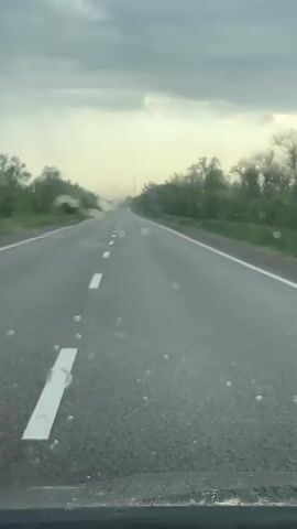 The video shows a view from inside a car, looking out through the windshield at a grey, overcast sky and a straight, empty road stretching ahead. The road has white lines marking the lanes and edges. On either side of the road, there's green vegetation, suggesting trees or bushes lining the roadside. Raindrops are visible on the windshield, slightly obscuring the view.  The wipers swipe across the windshield intermittently, clearing the water.