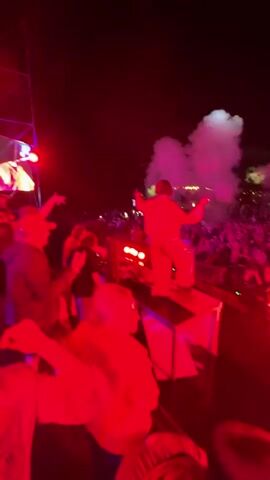 A crowd of people are gathered outdoors at night, seemingly at a concert or festival. The scene is illuminated by red and pink stage lighting. In the background, a large screen displays an image of a person. Several people in the foreground are dancing and holding up their phones. A person is visible on a raised platform, possibly a DJ booth, behind a console. Smoke or fog is visible in the air, illuminated by the lights. The overall atmosphere appears energetic and celebratory.