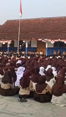 A large group of students, predominantly female and wearing brown hijabs and school uniforms, are sitting on the ground in rows. The students are facing a building with a red tiled roof and blue accents. A man in a white shirt and hat is standing on a raised platform in front of the building, speaking into a microphone. A flag, predominantly red and white, is flying from a flagpole to the left of the building. The students appear to be engaged in an assembly or outdoor class. The overall scene suggests an educational setting, possibly in Indonesia given the flag.