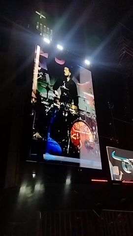 The video shows a concert performance being projected onto a large screen.  Initially, the screen displays a drummer and other musicians playing. The screen then flashes bright white and the video cuts out, leaving only the outline of the screen and nearby stage lighting visible.