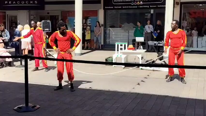 The video shows a street performance taking place outdoors in front of shops. Three performers, all wearing matching red and yellow outfits, are on a paved walkway. The performer on the right is holding a long white rope and appears to be interacting with it. The performer in the middle is standing with hands on hips, looking to the left. The performer on the left is facing away from the camera. A small audience of about a dozen people is gathered on the left side of the frame, watching the performance. In the background, shops with signs like "PEACOCKS" and "School Uniform" are visible. There is a keyboard and a white table with some props set up in the middle ground. The area is well-lit, suggesting it is daytime.
