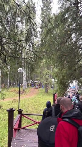 A group of people are walking in a line on a wooden walkway in a forest. The walkway has red railings. Some of the people are wearing backpacks. The trees are tall and green, and the ground is covered in grass. There are some small buildings or tents visible in the background. The sky appears to be overcast. The video is shaky, suggesting it was filmed while walking.