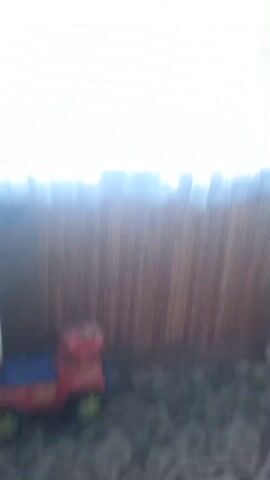 The video shows a room with wooden walls and a light-colored ceiling. There are windows with white curtains, letting in bright light. Various objects are visible, including a white appliance with a plastic bag on top, a shelf unit with items on it, a patterned rug, a circular braided rug, and a toy train. The camera is moving, and some parts of the video are blurry.