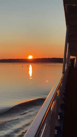 The video shows a sunset over a body of water as viewed from the deck of a ship. The sun is low on the horizon, casting a warm orange glow across the sky and reflecting on the water. The ship's railing is visible in the foreground, and the deck is made of wood. There are a few dark figures on the deck of the ship, and the water is rippling from the movement of the ship. The overall impression is one of tranquility and natural beauty.