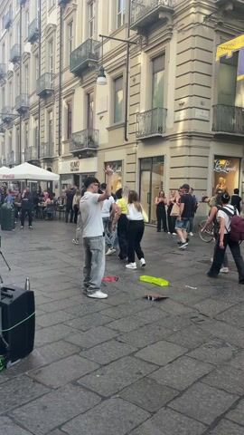 The video shows a young male street performer singing in a European city square. He is standing with a microphone in front of a black speaker, with a green donation basket in front of him. He wears a white t-shirt, loose light-colored jeans, and white sneakers. Pedestrians walk by him, and some seem to be listening to the performance. Some tables and chairs are visible at the edge of the square beneath a white tent. The buildings in the background are tall and ornate, with shops on the ground floor and balconies above. A sign on the corner building reads, “Via Roma, 31.” A man in a green shirt walks in front of the camera at the start of the video. The day is sunny and bright.