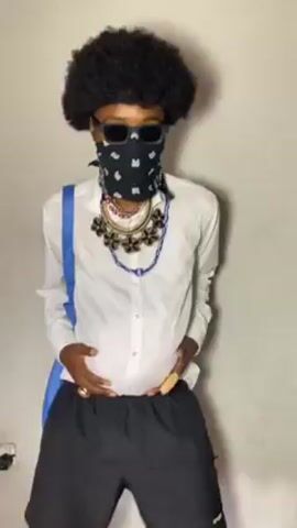 A man in an afro wig, sunglasses, and a bandana over his mouth is dancing in front of a plain grey wall. He is wearing a white dress shirt that appears too small for his stomach, along with loose black athletic shorts. He has multiple necklaces and is wearing rings on his fingers. A blue bag hangs over his shoulder. The man holds his stomach with both hands as he moves and dances to the music.