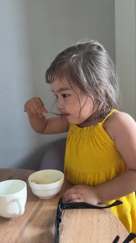 A young girl in a yellow dress sits at a wooden table, scooping food from a white bowl with a gold spoon. She places the spoon in a white cup and smiles at the camera. Another white bowl and sunglasses are also on the table. The background shows a light gray wall and part of a chair.