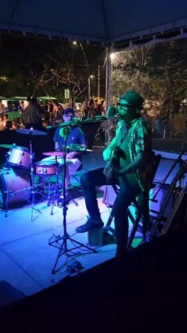 The video captures an outdoor musical performance at night in Armação dos Búzios, Brazil. Two musicians are featured on a small stage under a canopy structure.

On the right, a man with a hat and glasses is seated on a chair, playing an electric guitar and singing into a vintage-style microphone on a stand. He is wearing a patterned shirt and dark pants. The strong blue stage lighting illuminates him.

To the left, a drummer, also wearing glasses, is playing a drum kit. The drum kit includes a bass drum, snare drum, and cymbals. The bass drum has the text "D'Addario" visible on it. Pink and purple lighting highlights the drummer and his instrument.

In the background, a crowd of people can be seen milling around, indicating a public event or gathering. Trees and ambient street lighting are also visible beyond the performance area. The ground under the stage appears to be paved. The overall scene suggests a lively evening entertainment event.