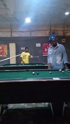 The video shows two men playing pool in what appears to be a recreational establishment, possibly a bar. The man on the right wears a denim shirt and a blue baseball cap. He is standing by a pool table, gesturing with his hands. On the table in front of him are several pool balls. The second man, wearing a yellow shirt, is across the room at another pool table. He is aiming his pool cue at the balls on the table. There are posters on the walls behind them, and the room is illuminated by overhead lighting. The room has a casual and somewhat aged appearance.