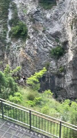 A person in a harness is seen swinging back and forth on a long rope over a deep canyon, with rocky cliffs on either side. Below the person, there is a lush green valley with trees and shrubs. Above the person, there are two metal bridges, one of which has people walking on it. The camera is positioned on a lower metal walkway, looking up at the swing and the canyon.