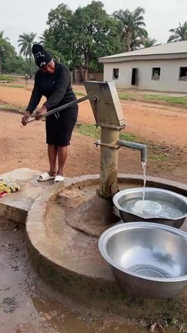 A woman in a black outfit and white sandals stands on a concrete platform and operates a hand water pump. Water flows from the pump into a metal bowl on the platform. Another empty metal bowl sits below it. The platform is surrounded by red dirt. In the background, there is a building, trees and palm trees.